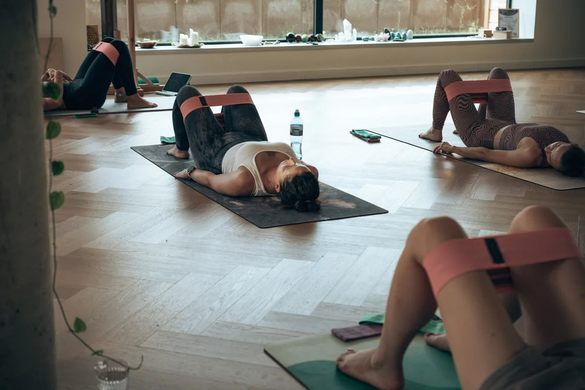 Uczestniczki zajęć pilatesu leżące na matach z gumami oporowymi na udach, w jasnej sali z drewnianą podłogą – pilates camp